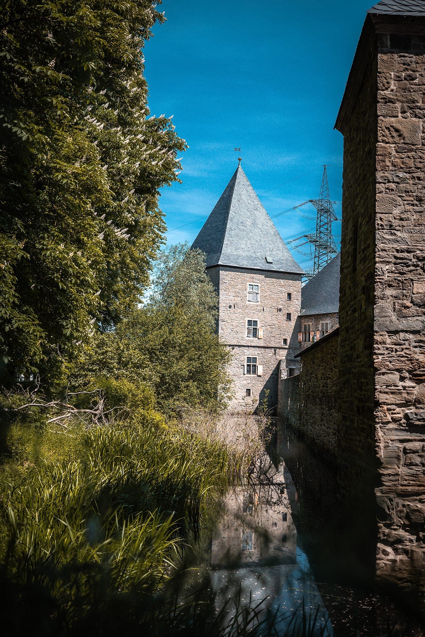 Der schiefergedeckte Turm von Haus Kemnade hinter einer blühenden Kastanie, gespiegelt im Wassergraben