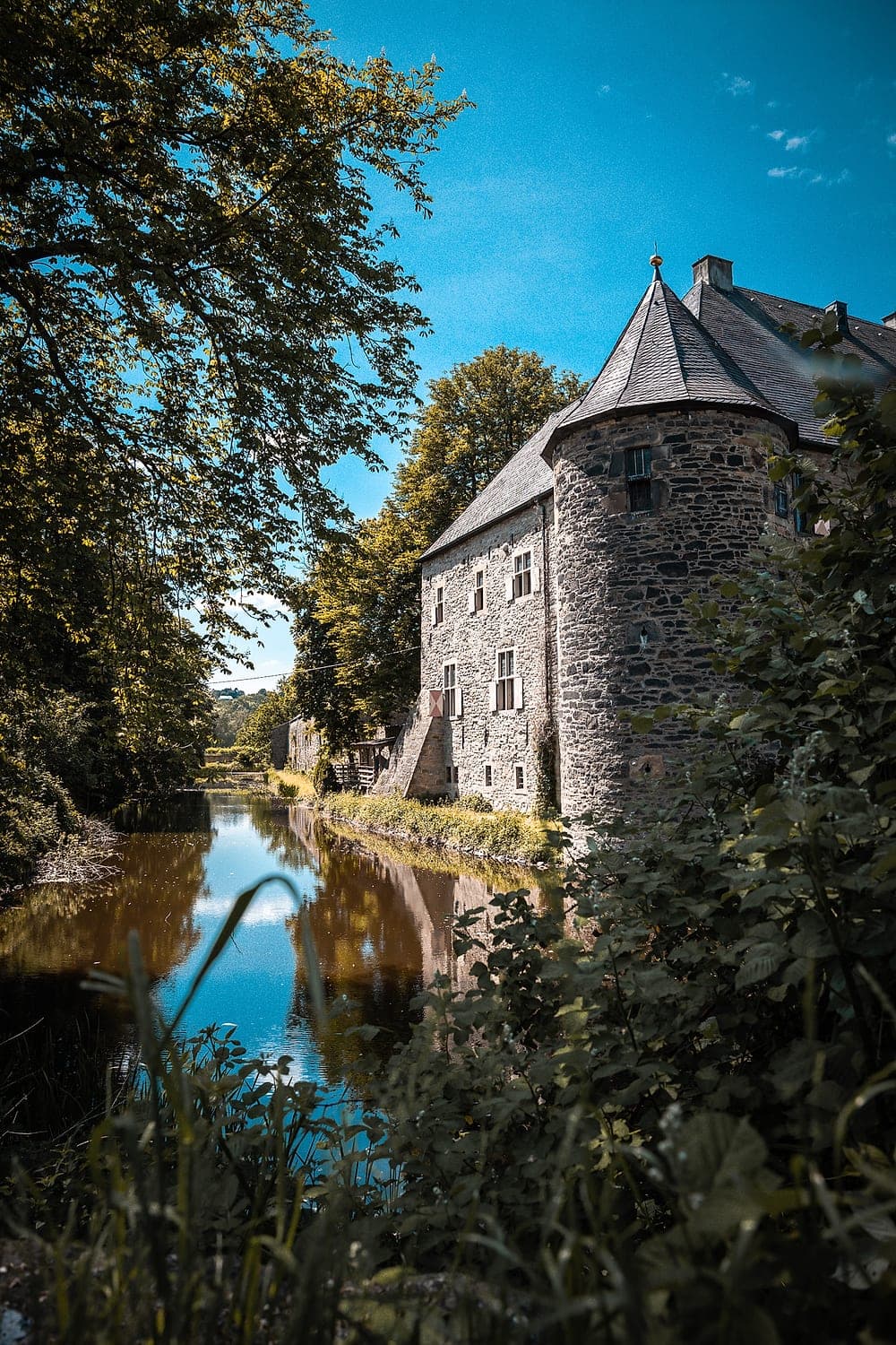 Der Rundturm der Wasserburg über dem Wassergraben, umrahmt von alten Bäumen