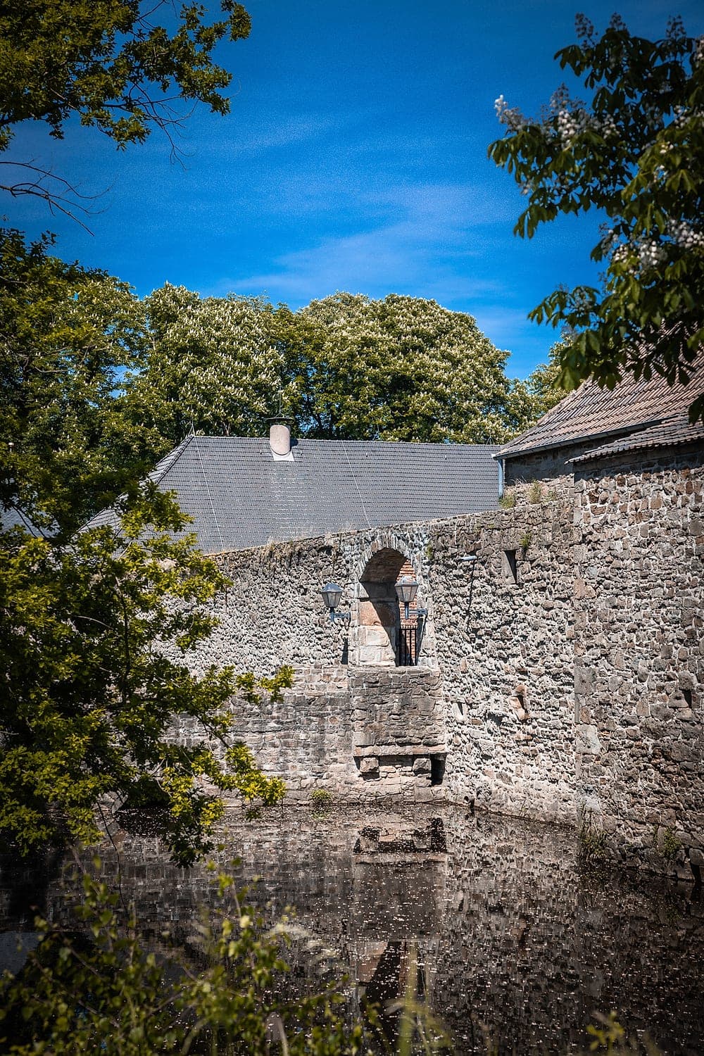 Torbogen mit Laternen in der Bruchsteinmauer, davor der Wassergraben, dahinter blühende Kastanien