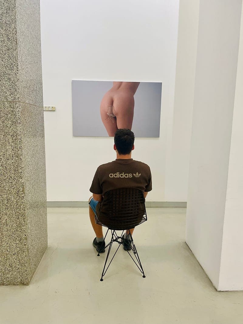 Hendrik sitzt in einer Galerie auf einem Drahtstuhl vor einem großformatigen Foto